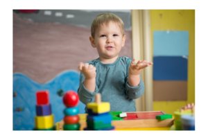 Boy standing at table with nesting toys holding hands up in air with questioning look