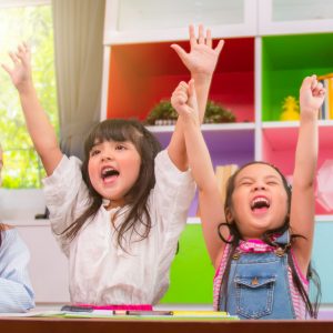 Two girls with both hands raised and shouting in approval in a classroom with brightly colored cubbies.