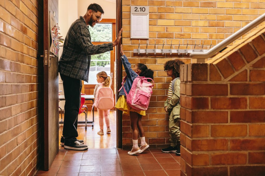 Preschool teacher greeting his students at the door. Male teacher welcoming his class with a high five